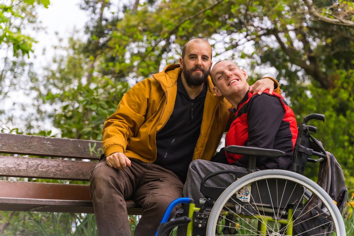 A smiling young man in a wheelchair with a friend's arm around his shoulder, sitting by a park bench