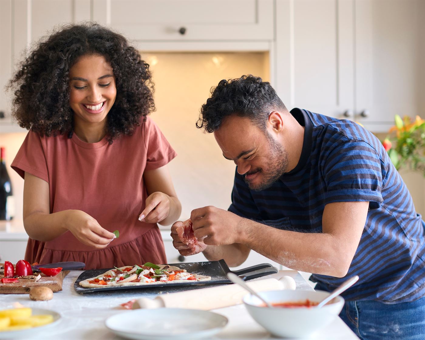 A young man and a young woman laughing as they make a pizza together in a home kitchen